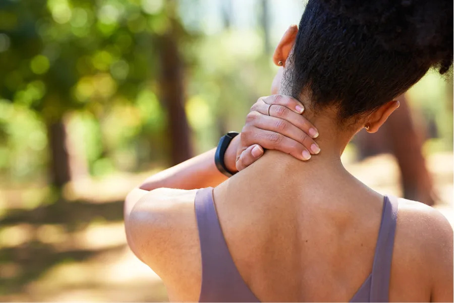 Woman outdoors holding her neck in pain, wearing a sleeveless top with blurred natural background