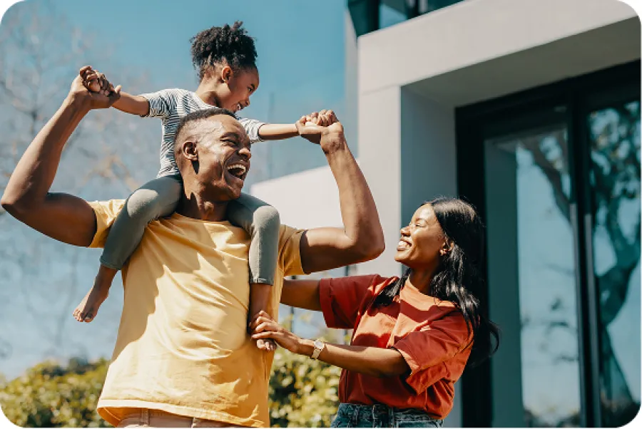 Happy family outdoors with father carrying daughter on shoulders and mother smiling beside them.