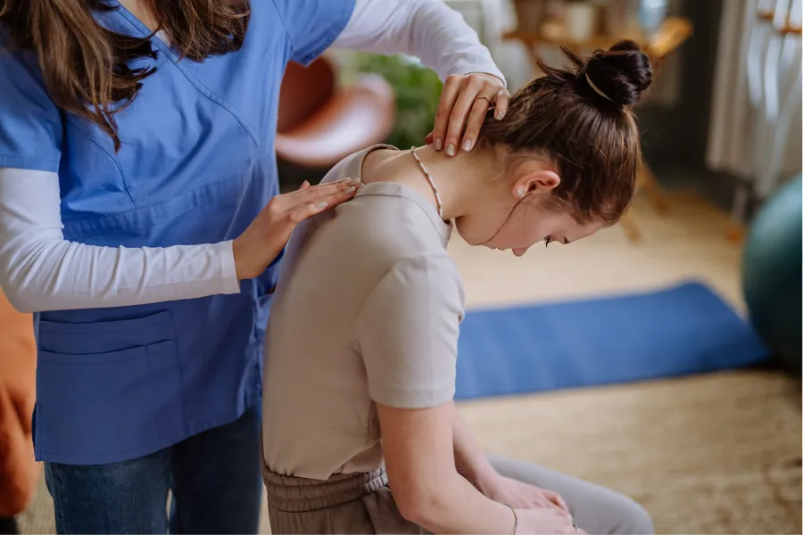 Therapist in blue scrubs performing neck adjustment on seated woman during physical therapy session.