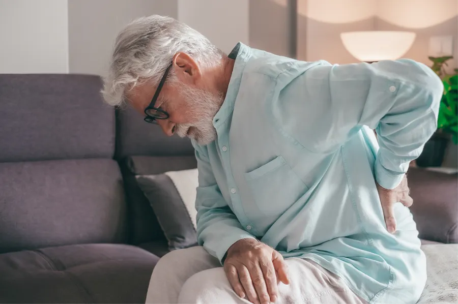 Elderly man sitting on couch holding lower back in pain wearing glasses and light blue shirt indoors.