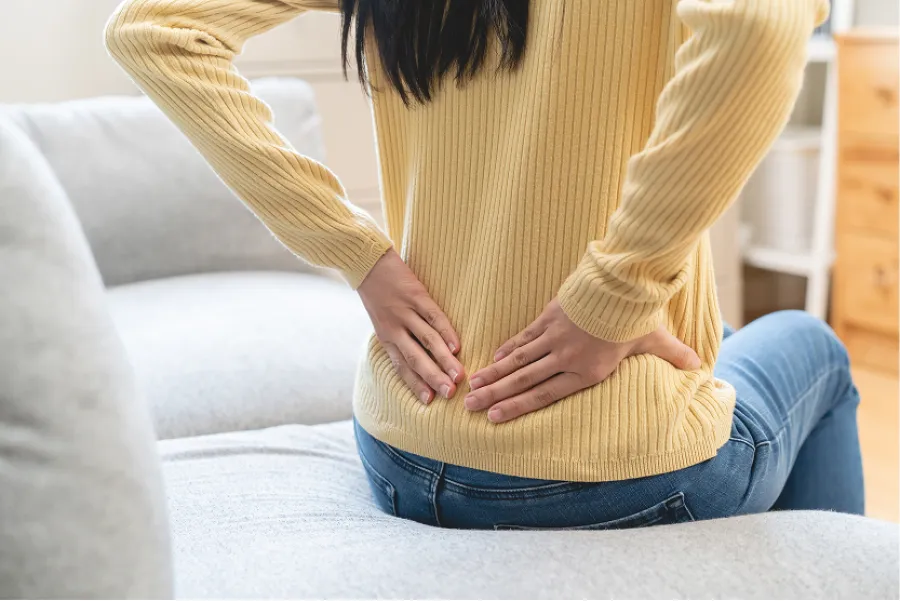 Person wearing yellow sweater sitting on a couch holding lower back in pain or discomfort indoors.