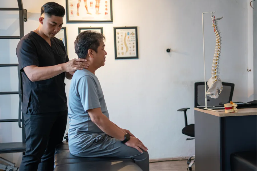 Chiropractor performing neck adjustment on seated middle-aged man in clinical setting with spine model and charts.