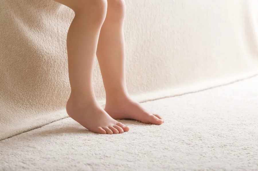 Close-up of a child's bare feet and lower legs on a soft beige carpet near a textured wall.