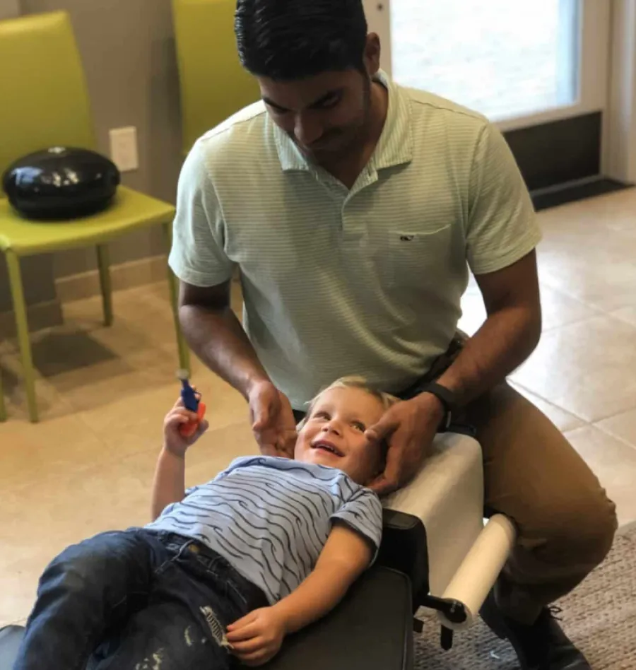 Child smiling while receiving a gentle chiropractic adjustment from a male therapist in a bright clinic room.