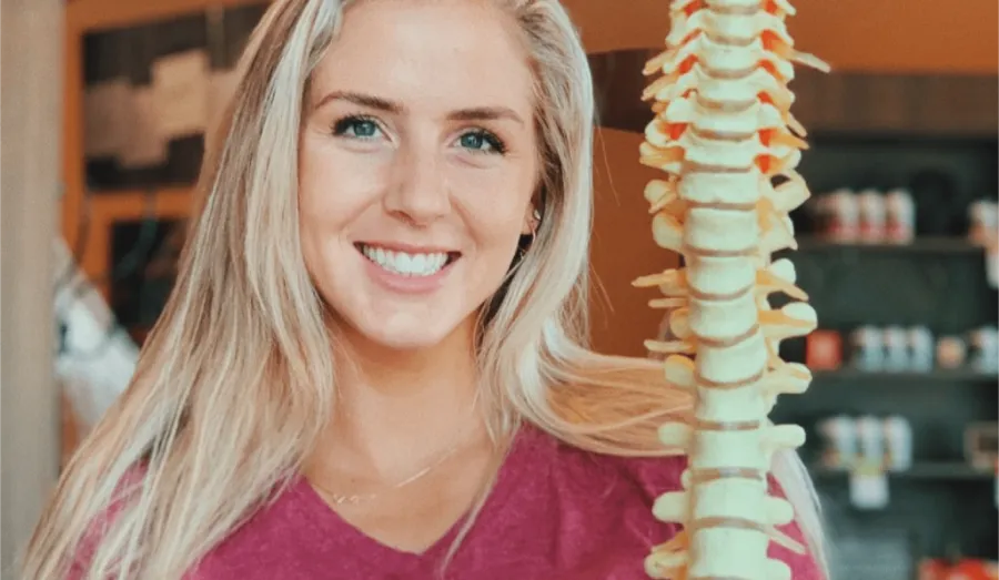 Smiling woman with long blonde hair holding a model of the human spine in a medical setting.