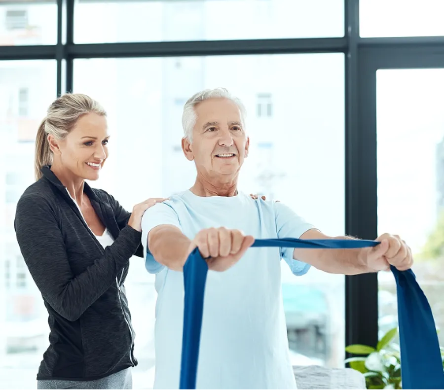 Senior man exercising with resistance band assisted by a female physical therapist in a bright room.
