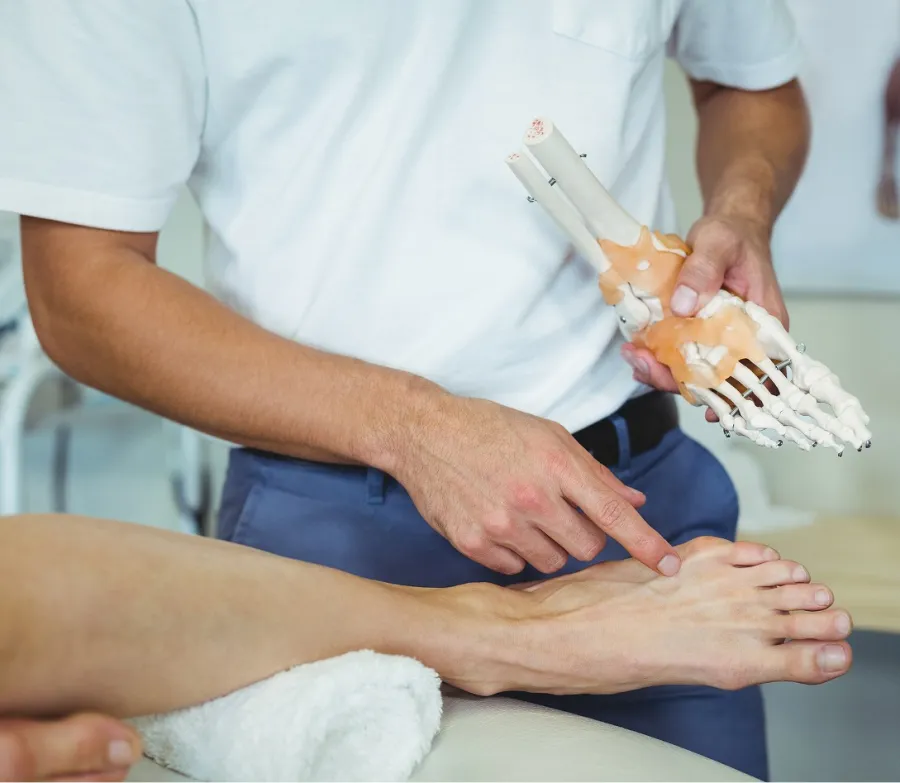 Doctor explaining foot anatomy using a skeletal model while pointing at a patient's foot in a clinical setting.
