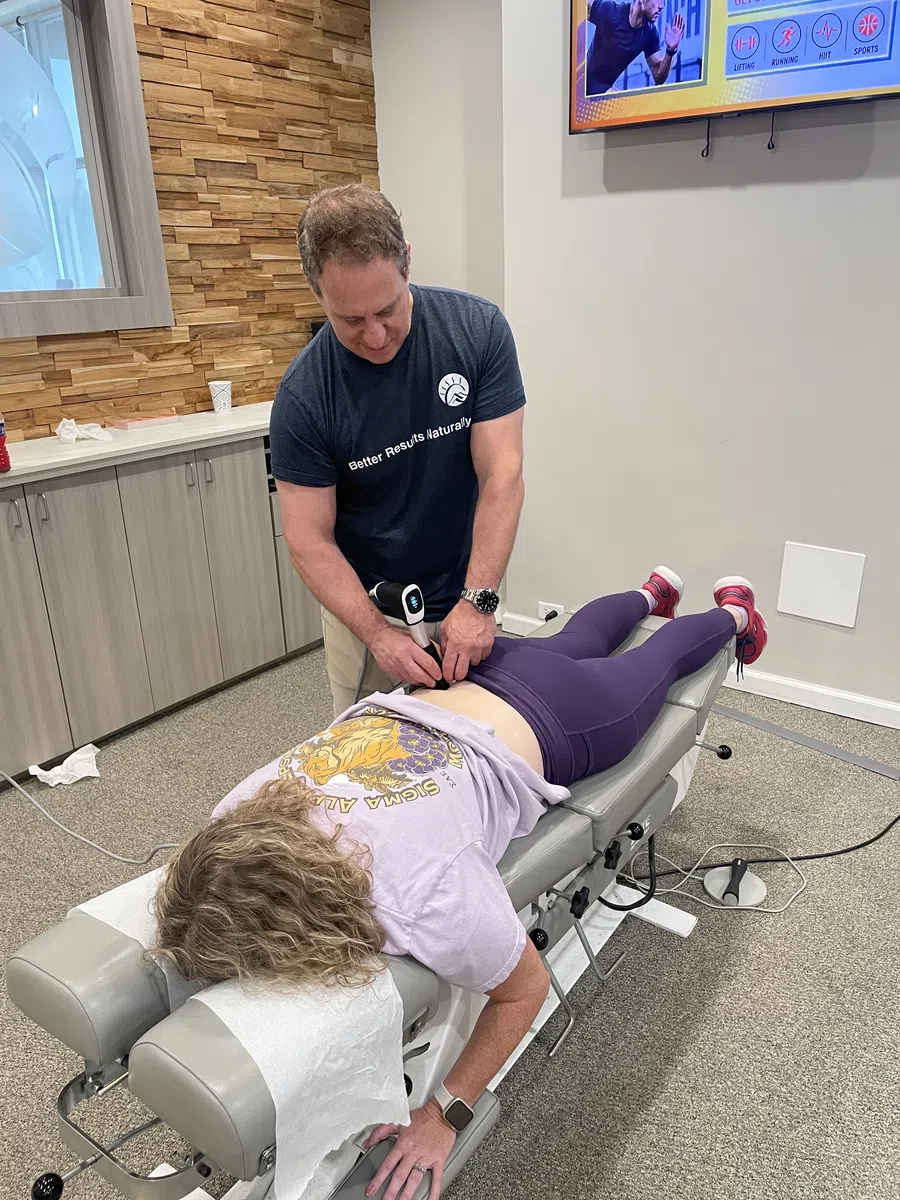 Chiropractor applying therapy device on patient's lower back on treatment table in clinic setting.