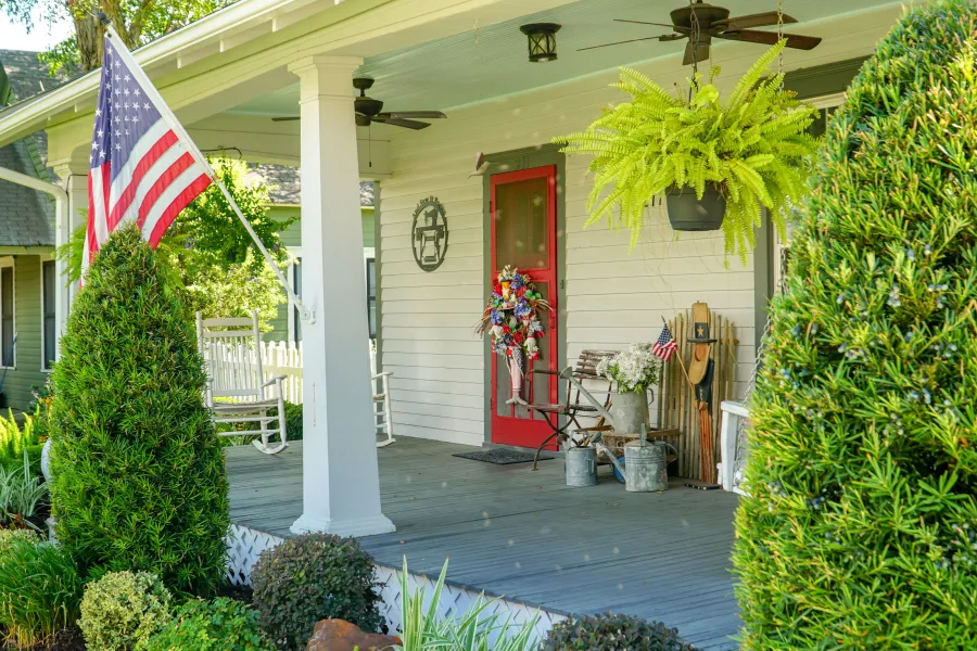 Cozy front porch with red door, American flag, rocking chairs, hanging fern, and decorative plants