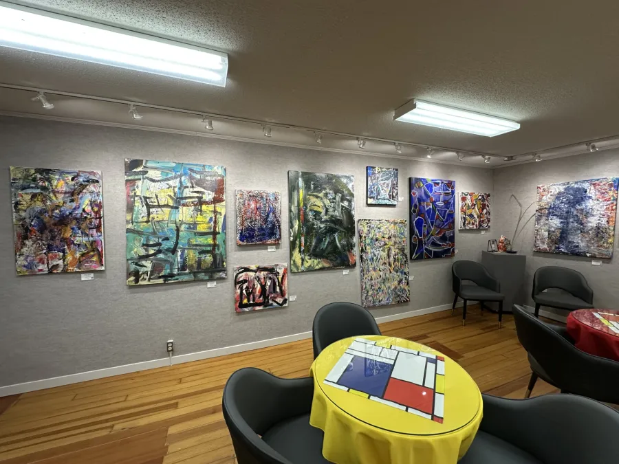 Contemporary abstract paintings displayed on a gallery wall with chairs and tables in the foreground.