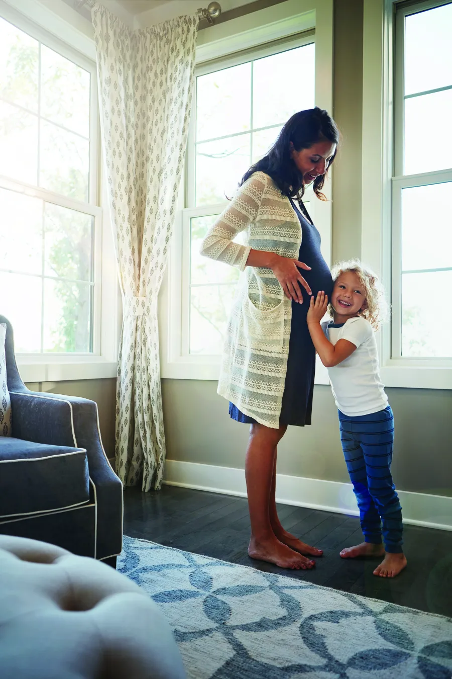 Pregnant woman standing by windows with child smiling and hugging her belly in a cozy living room