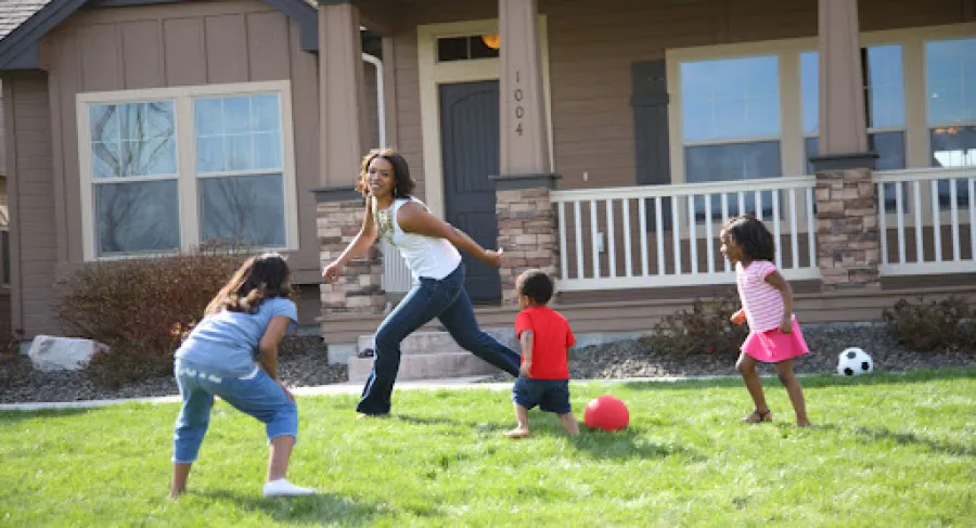 A woman playing with children in a green yard outside a suburban home, enjoying a sunny day together.