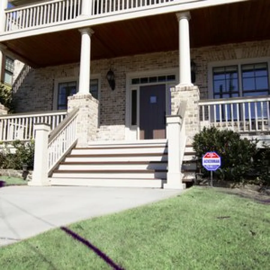 Front porch of brick house with wooden columns, steps, and security sign near bushes