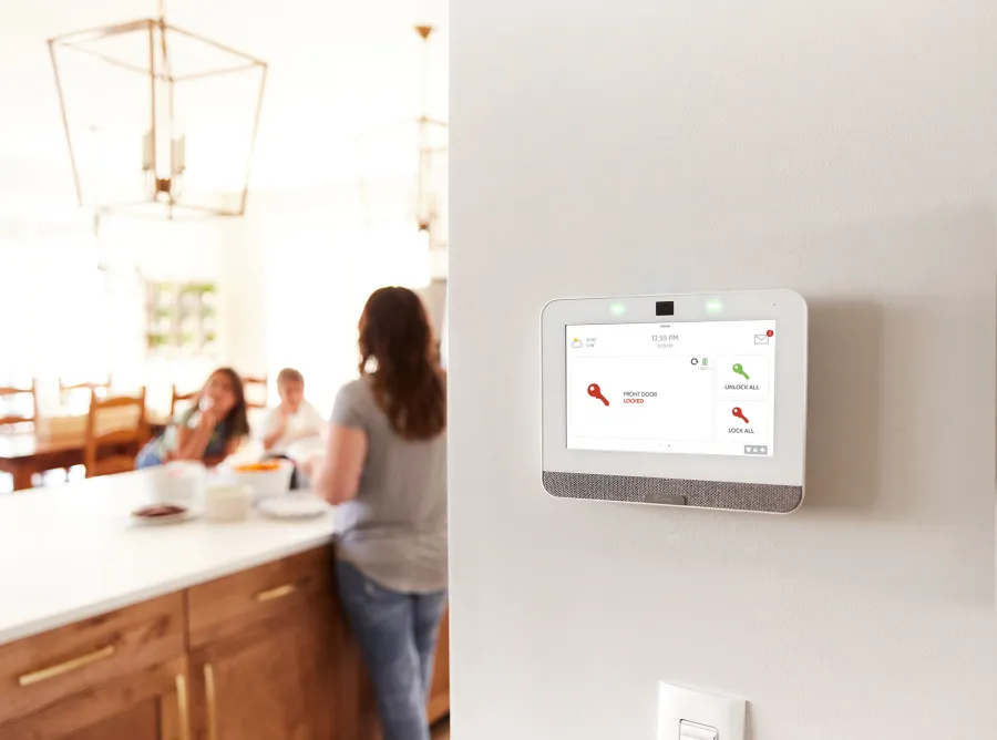 Smart home security touchscreen panel on wall showing front door locked and options in a bright modern kitchen.