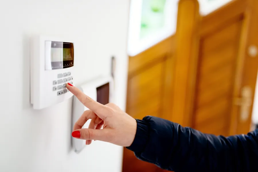 Woman with red nail polish entering code on a white home security alarm keypad on a wall.