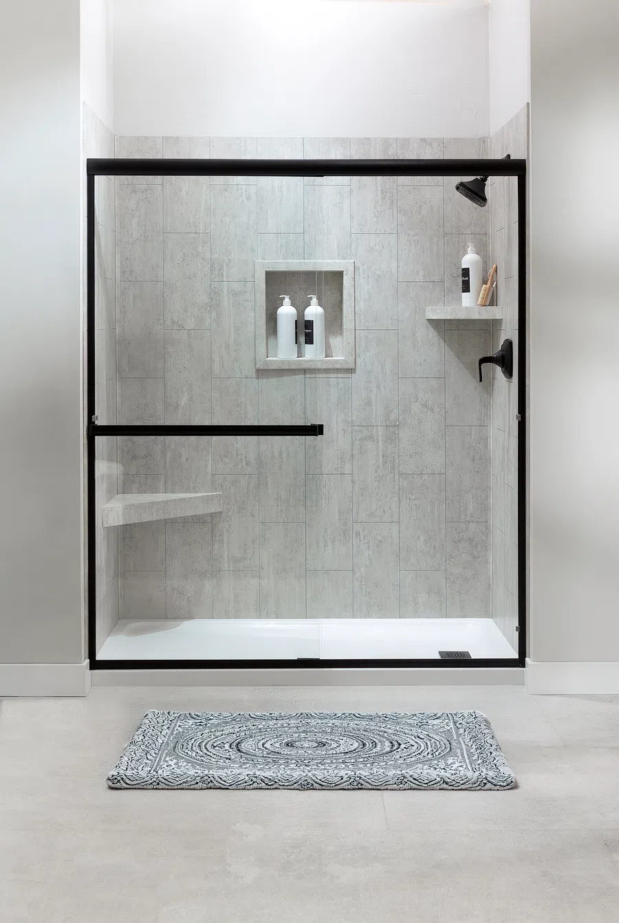 Modern walk-in shower with glass doors, gray tile walls, built-in shelves, and patterned bath mat
