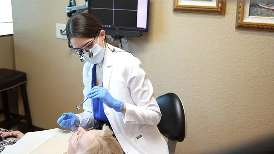 Female dentist in a white coat and mask examining an elderly patient's teeth in a dental clinic.
