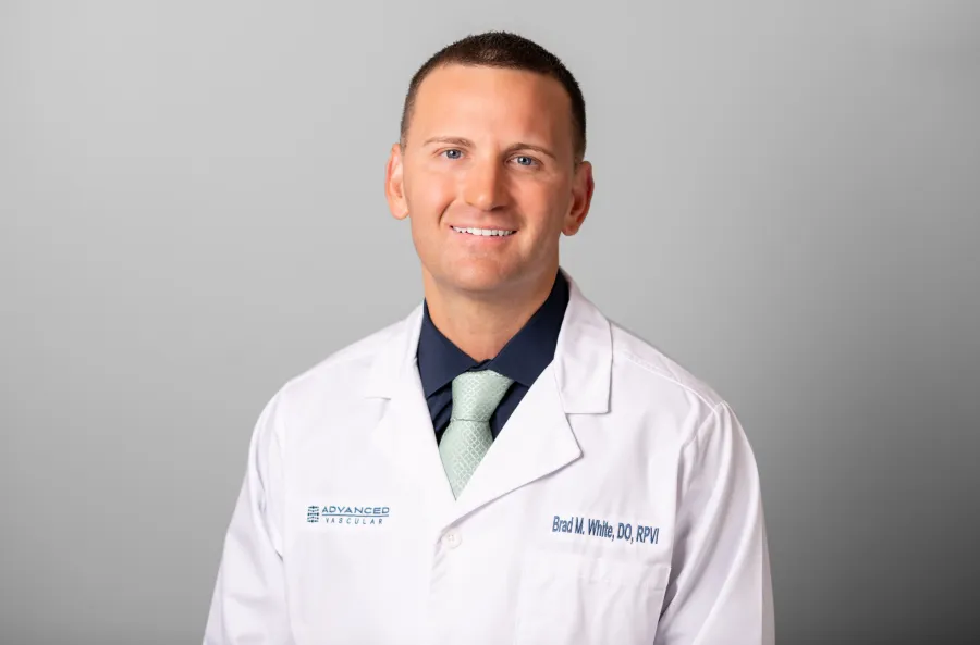 Smiling male doctor in white coat with green tie on gray background, representing advanced vascular care.