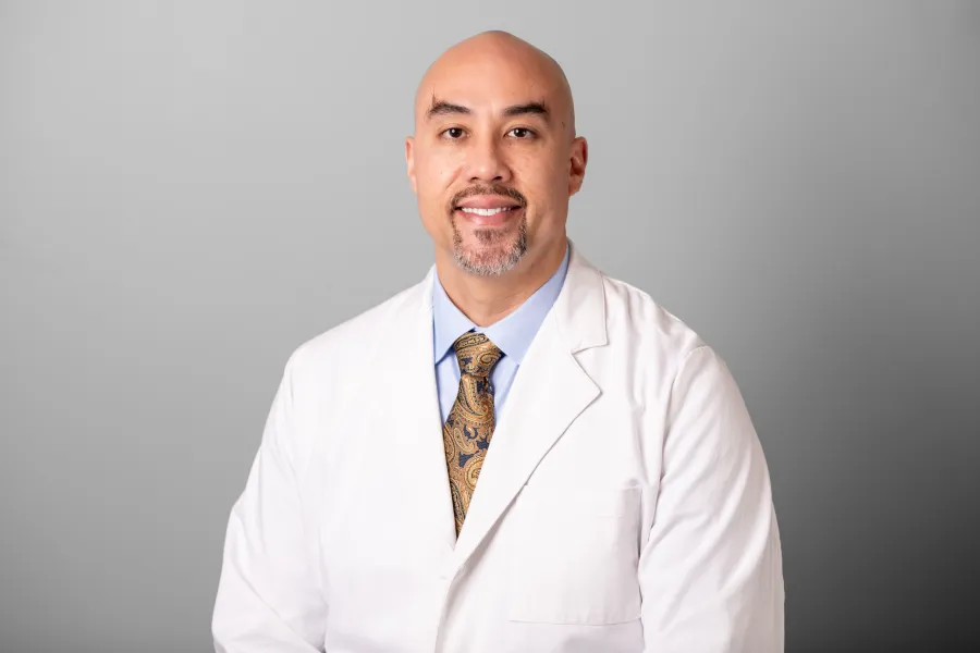 Portrait of a smiling male doctor wearing a white coat, blue shirt, and patterned tie against a gray background