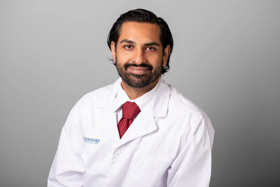 Smiling male doctor in white lab coat and red tie against plain gray background.
