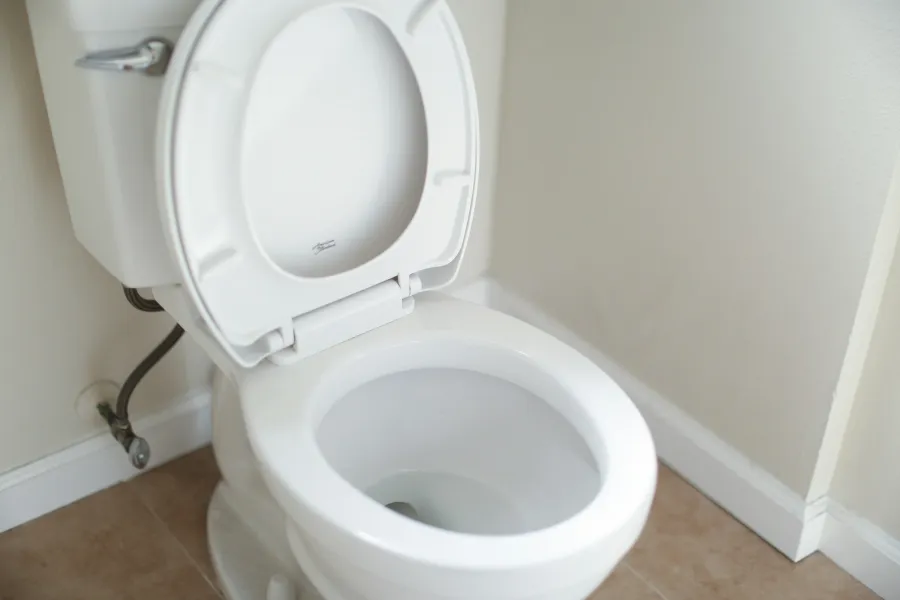 Clean white toilet bowl with open lid in a beige tiled bathroom corner with white walls.