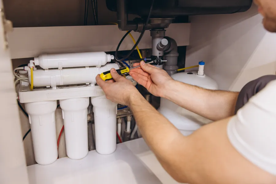 Technician repairing or installing a water filtration system under a kitchen sink using a screwdriver.