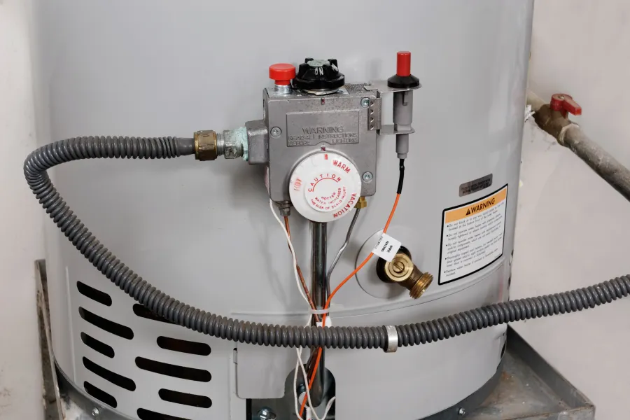 Close-up of a water heater temperature control knob and connected pipes with warning label visible.