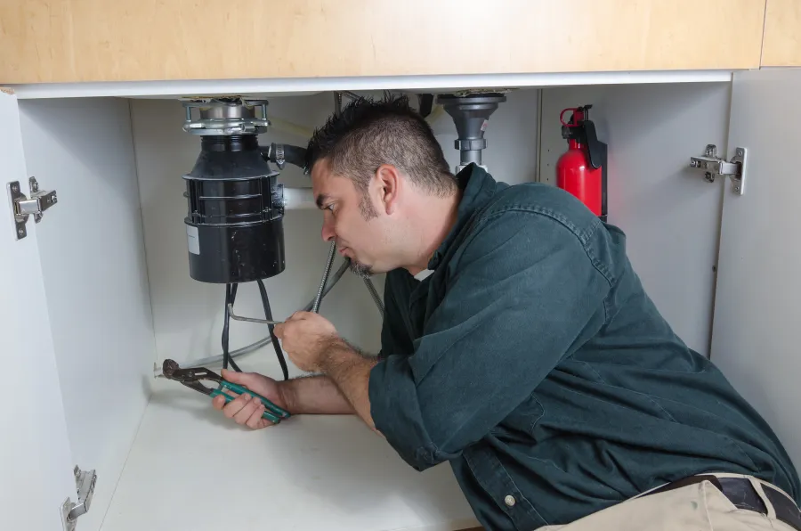 Plumber repairing kitchen sink drain using adjustable wrench under the cabinet next to a fire extinguisher.