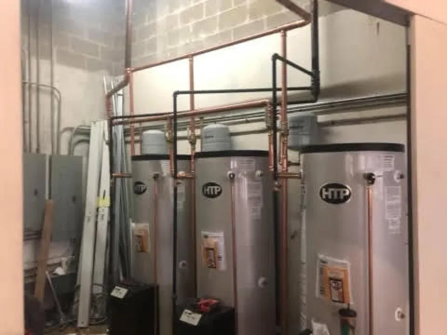 Three large HTP water heaters connected with copper pipes in a utility room with concrete walls.