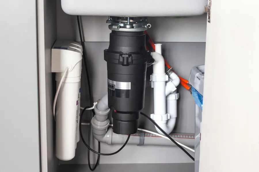 Under-sink view showing a garbage disposal unit, water filter, and connected plumbing pipes.