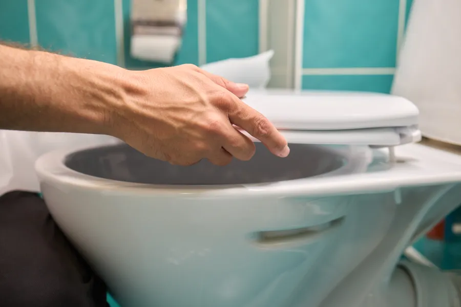Hand lifting the lid of a white toilet bowl in a bathroom with teal tiles and toilet paper holder.