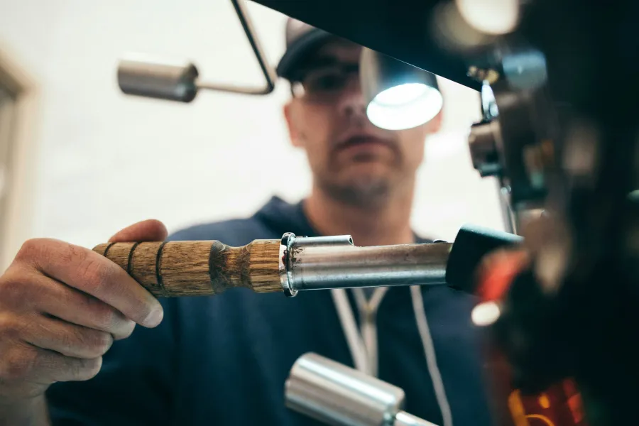 Barista tamping coffee grounds with wooden handled tool in espresso machine portafilter during brewing.