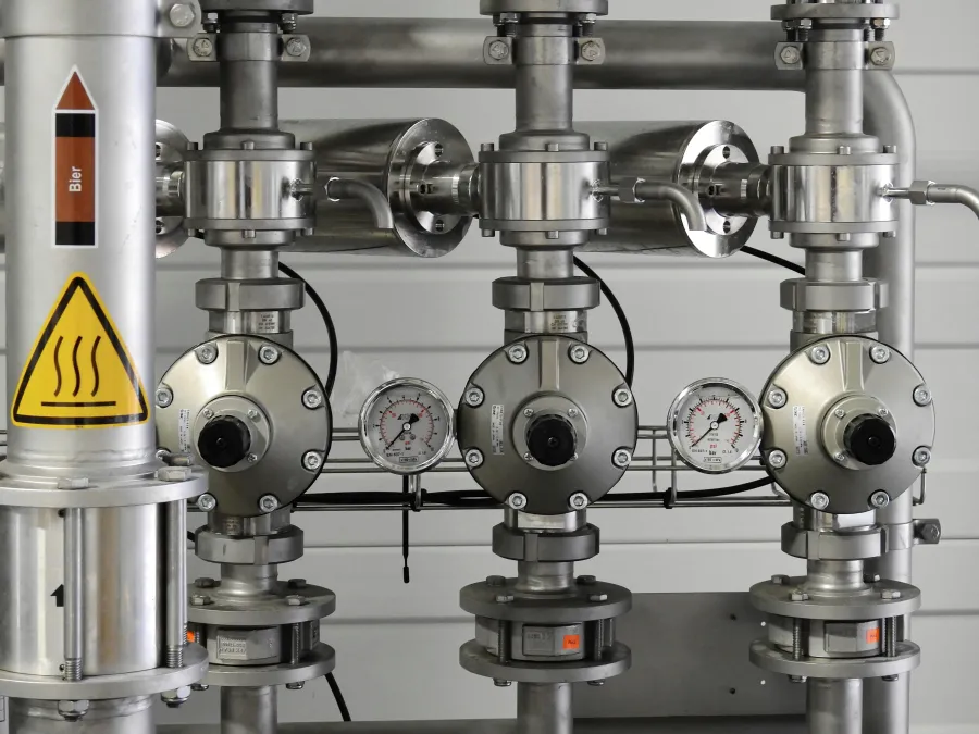 Close-up of industrial stainless steel pipes and pressure gauges in a brewing system with warning labels visible.