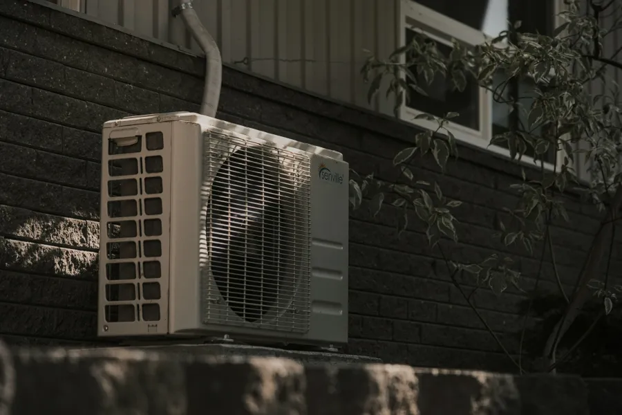 Outdoor air conditioning unit mounted on a dark brick wall with foliage nearby in soft lighting