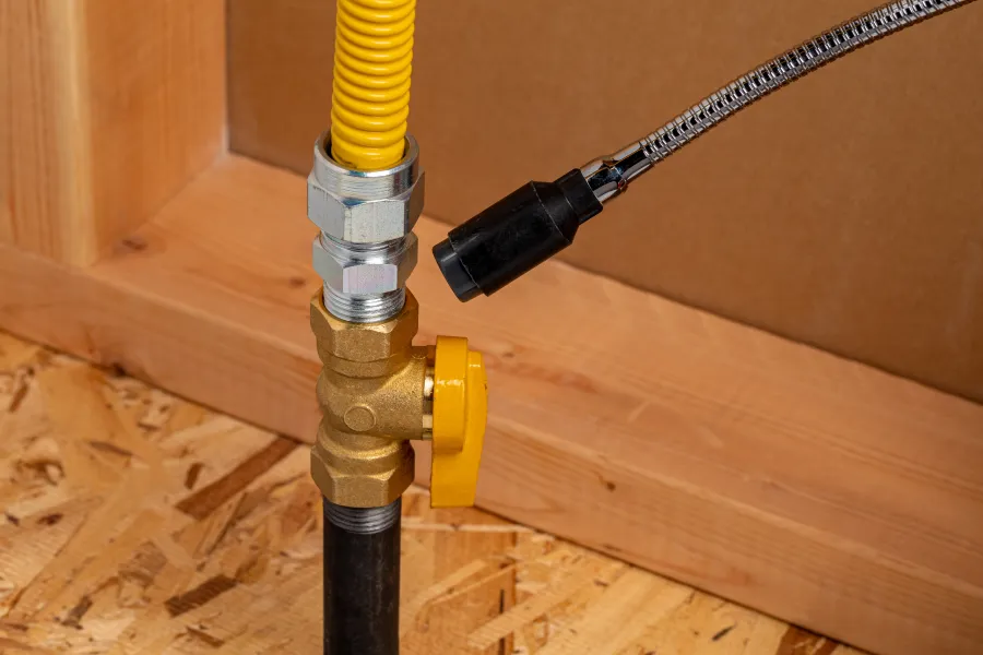 Close-up of a yellow gas valve and flexible connector pipe installed on a wooden floor and wall framing.