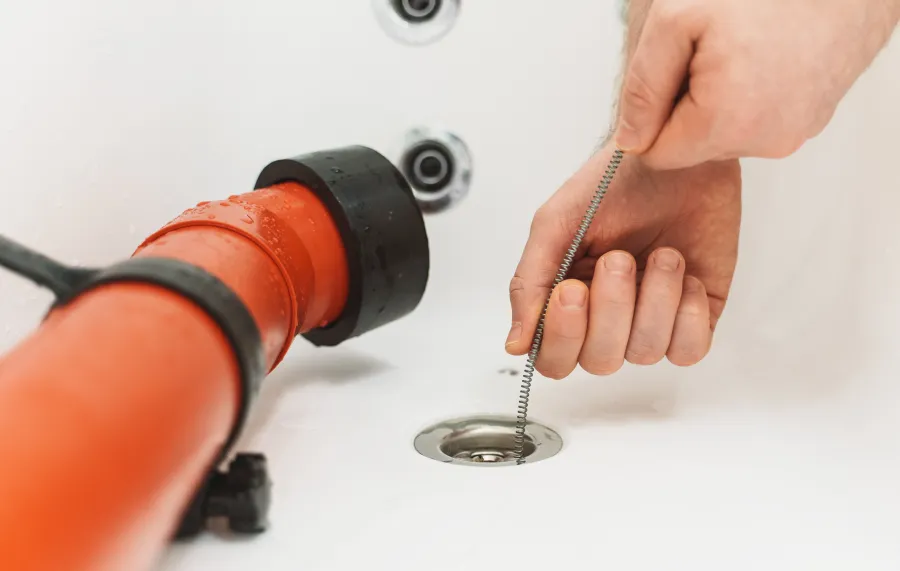Hand using drain snake tool to clear clogged bathtub drain near red plumbing pipe and faucet.