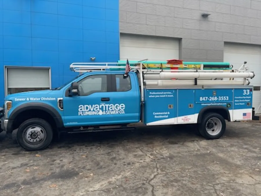 Blue Advantage Plumbing service truck with ladders and tools parked outside a building with blue and gray walls.