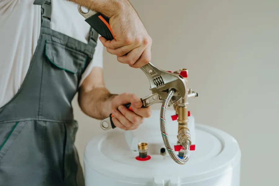 Plumber using adjustable wrenches to connect a braided hose to a water heater valve in a white boiler.