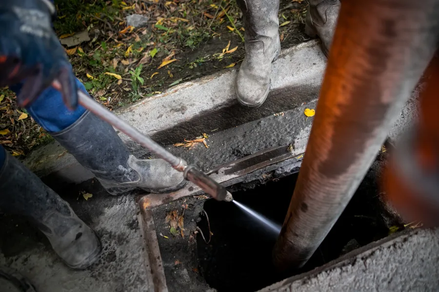 Workers wearing boots use a high-pressure water jet to clean a storm drain outside on a cloudy day