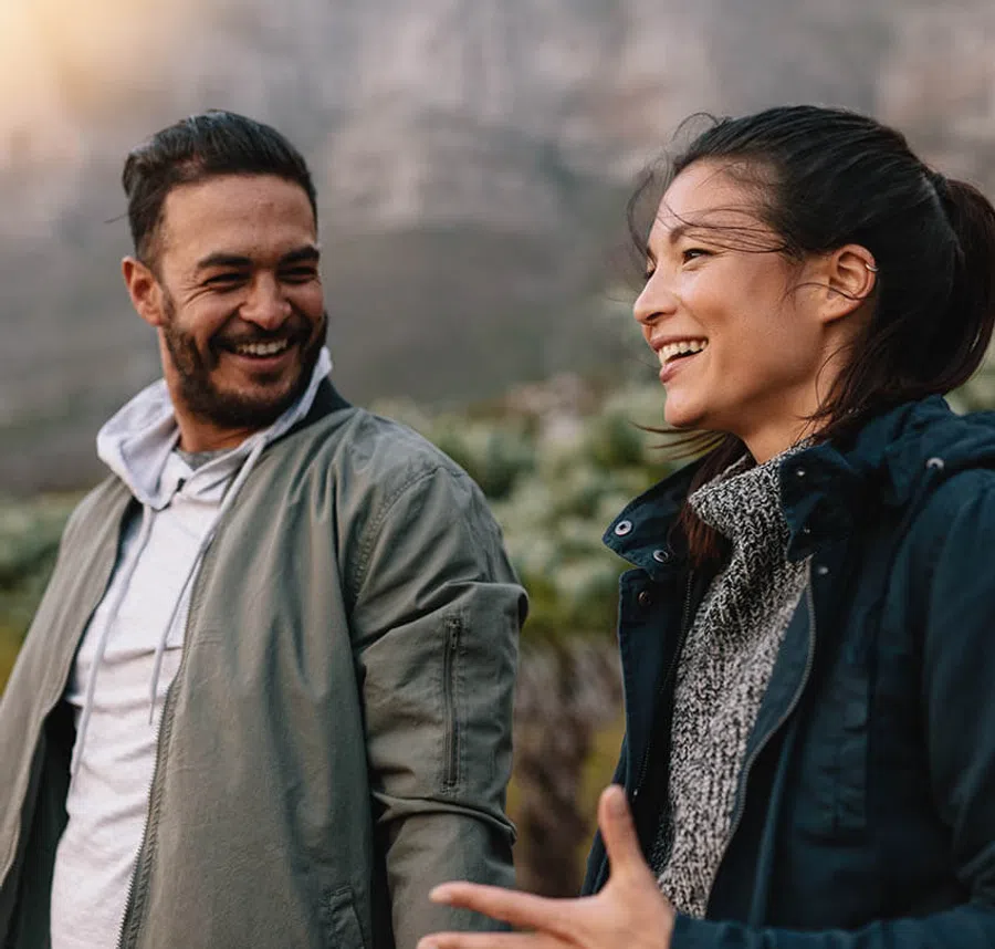 Smiling man and woman outdoors in casual jackets enjoying a conversation with blurred nature background.
