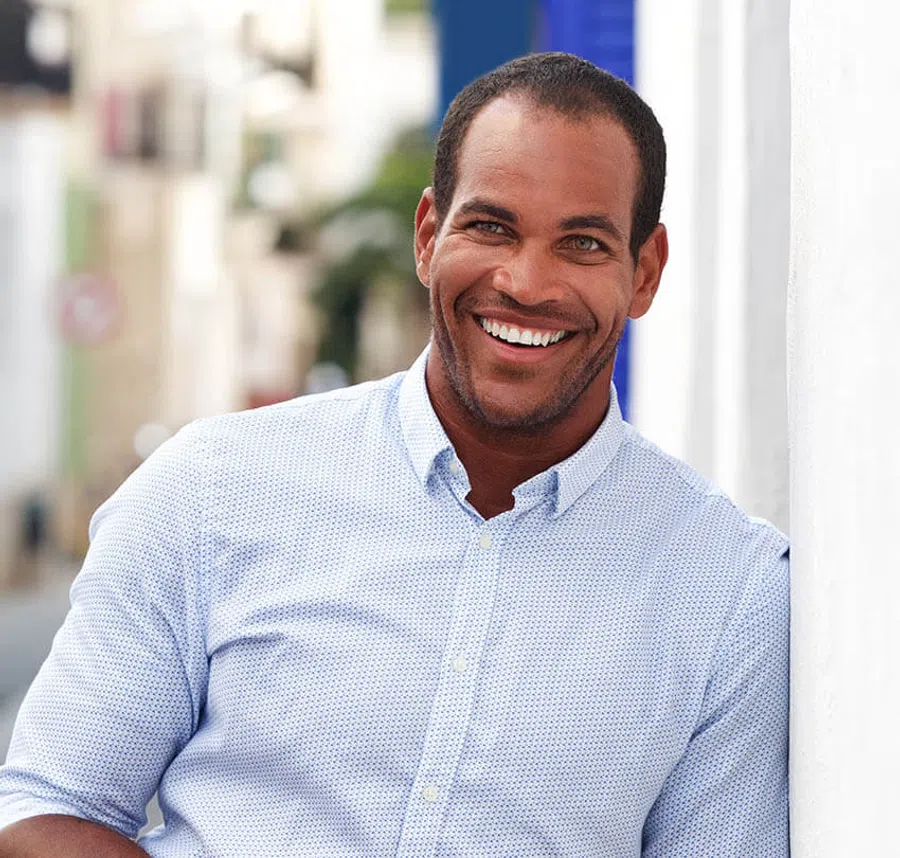 Smiling man in light blue patterned shirt leaning against a white wall in a bright urban setting.