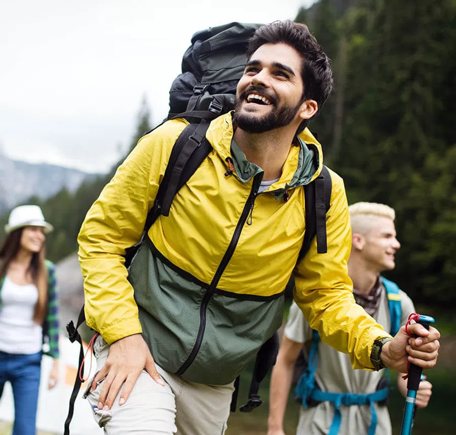 Smiling man in yellow jacket hiking with backpack and trekking pole in a forested mountain area.