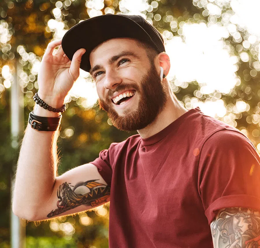 Smiling bearded man in a maroon shirt and black cap with tattoos, enjoying sunny outdoor setting.