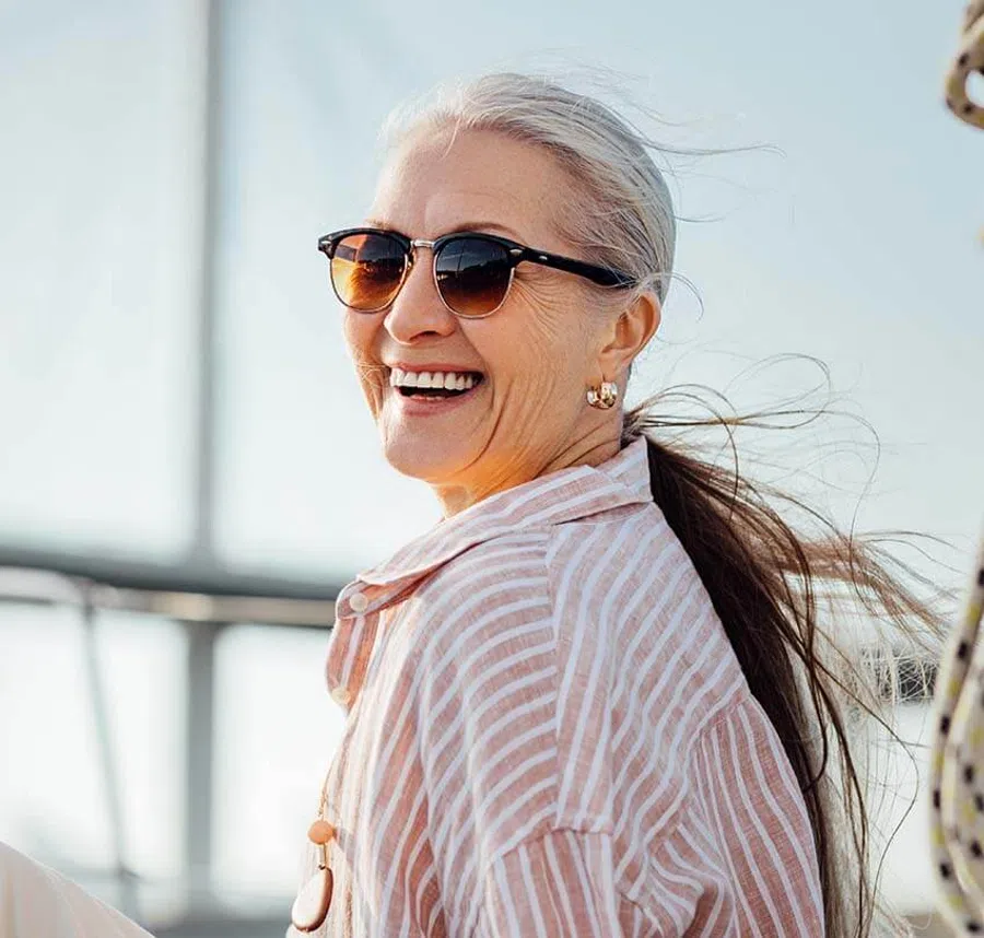 Smiling older woman with gray hair in ponytail wearing sunglasses and a striped shirt outdoors in sunlight.