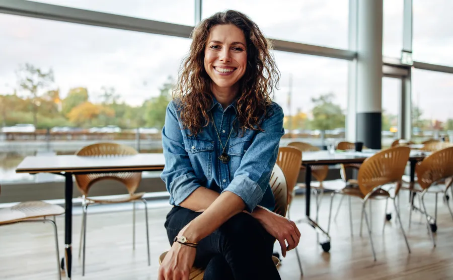 Smiling woman in denim shirt sitting cross-legged in bright modern room with wooden chairs and tables.