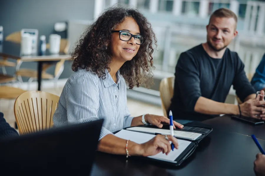 Smiling woman in glasses taking notes during a business meeting with colleagues in a modern office.