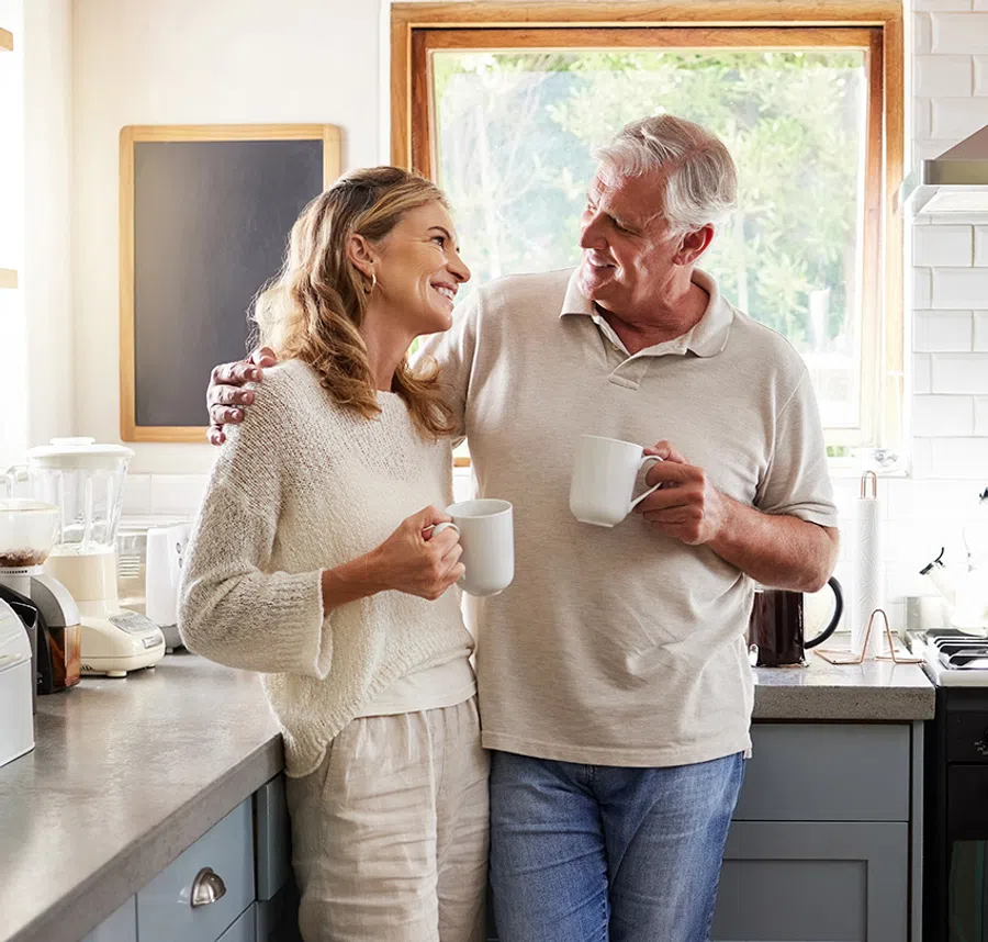 Happy mature couple smiling and holding mugs while enjoying coffee in bright modern kitchen