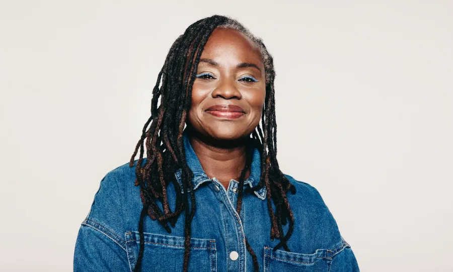 Smiling woman with long dreadlocks wearing a blue denim jacket against a plain light background.