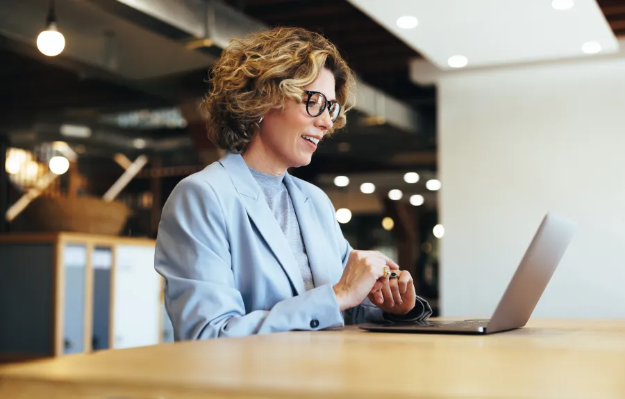 Smiling woman with glasses working on laptop in modern office setting with warm lighting.