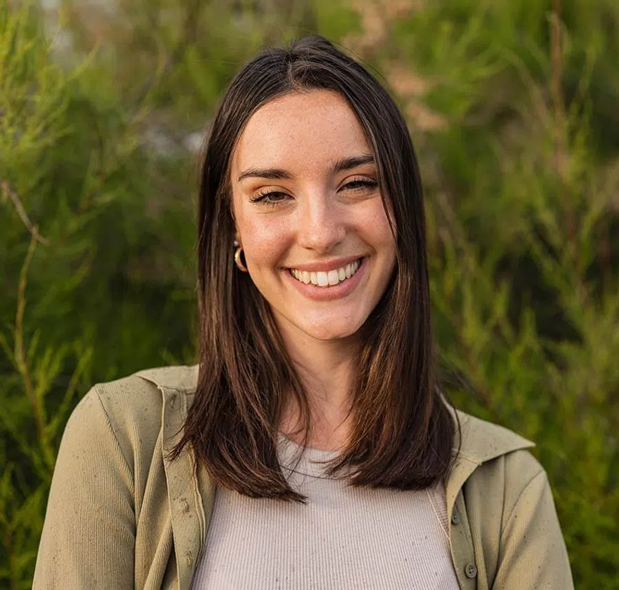 Smiling young woman with straight brown hair outdoors in casual beige and olive clothing with greenery background